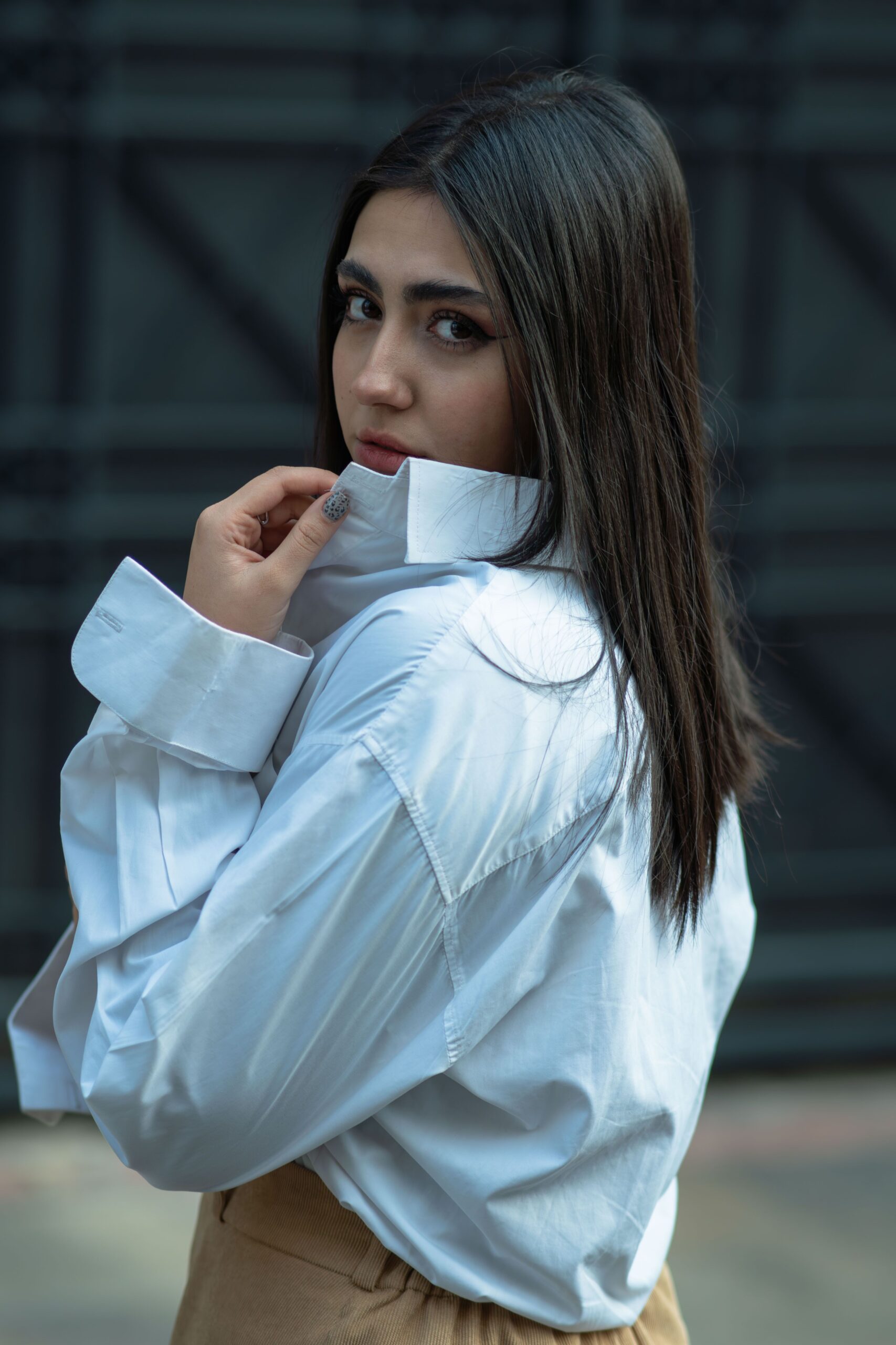 Chic woman with long hair poses confidently in a white shirt outdoors.